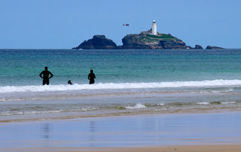 Godrevy Helicopter This landscape photograph captures the Godrevy helicopter flying near the iconic Godrevy Lighthouse, which is located off the coast of Cornwall, during a late morning in the summer season. The image shows the turquoise sea and the sandy beach in the foreground, where several people can be seen wading and swimming in the gentle surf. The lighthouse stands prominently atop Godrevy Island, providing a well-known landmark that is often associated with the coastline of Cornwall. The calm sea and bright beach under clear skies emphasize the coastal character of the region, highlighting the connection between the beach, the sea, and the historic Godrevy Lighthouse.